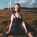 Sustainable Fashion. - Side view of graceful young female ballet dancer in elegant dress and pointe shoes sitting on old stone fence with closed eyes near windmills in countryside