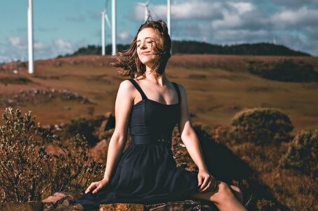 Sustainable Fashion. - Side view of graceful young female ballet dancer in elegant dress and pointe shoes sitting on old stone fence with closed eyes near windmills in countryside