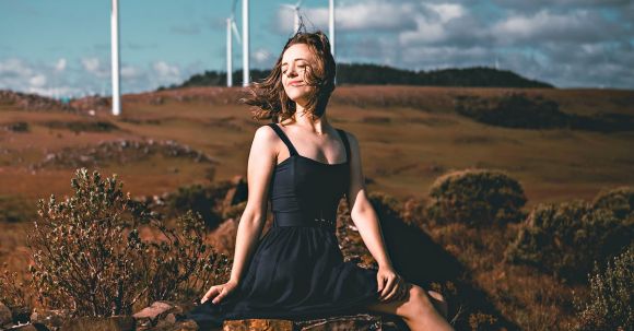 Sustainable Fashion. - Side view of graceful young female ballet dancer in elegant dress and pointe shoes sitting on old stone fence with closed eyes near windmills in countryside