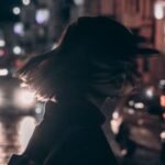 Fashion Movement - Woman in Black Jacket Carrying Backpack With Tousled Hair Walking On The Street At Night