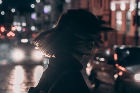 Fashion Movement - Woman in Black Jacket Carrying Backpack With Tousled Hair Walking On The Street At Night