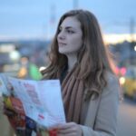 Clothing Exchange Guide - Female traveler in warm clothes standing on street with map and looking away at dusk